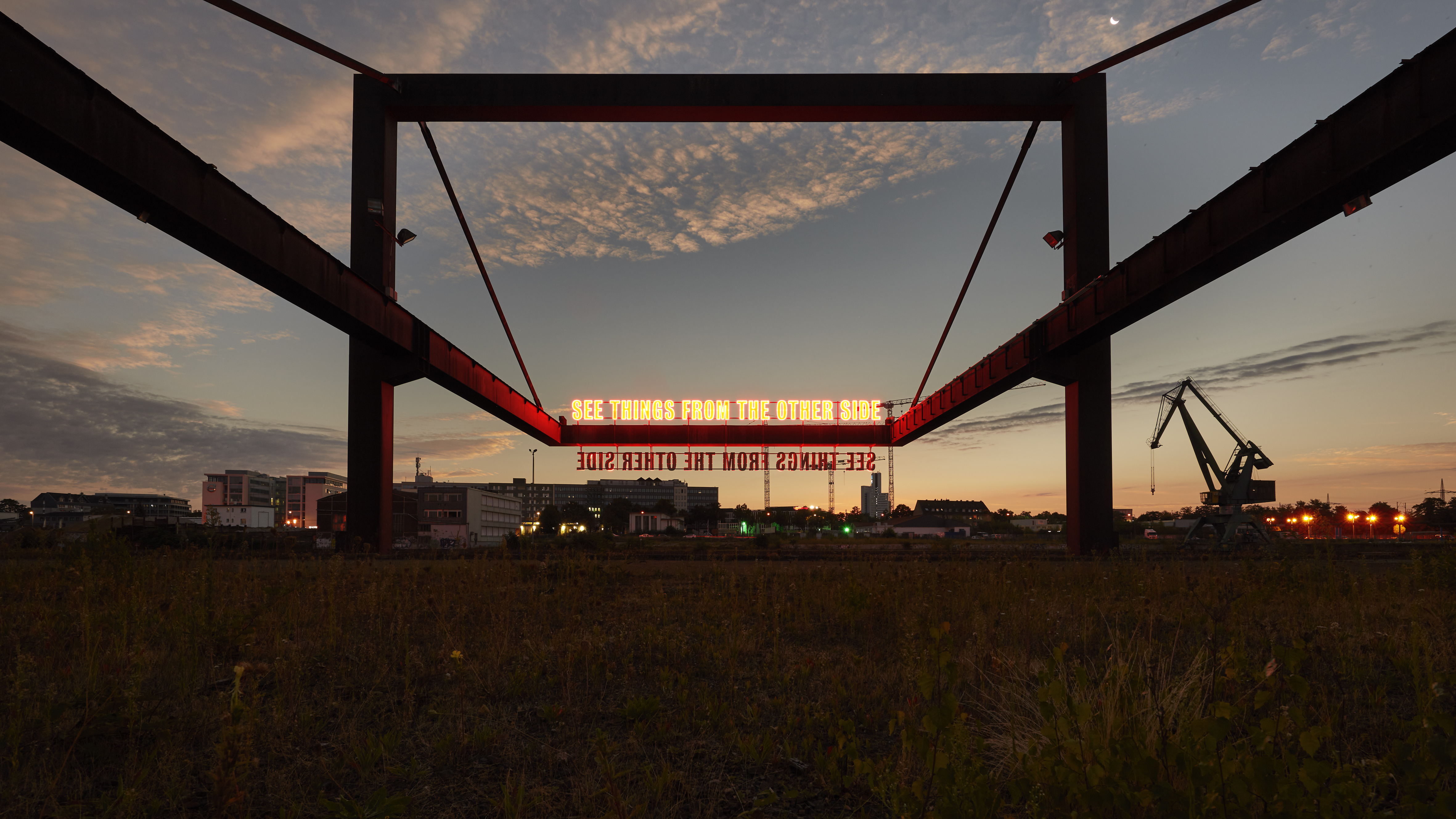 Lichtinstallation von Tim Etchells im Deutzer Hafen bei Dämmerung: rot leuchtender Schriftzug „SEE THINGS FROM THE OTHER SIDE" in einem Stahlrahmen, dahinter Hafenkran und Stadtsilhouette